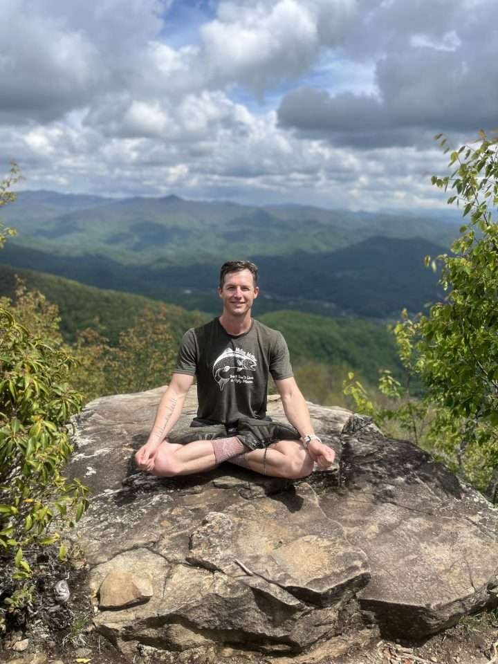 Zach Mills in Lotus Pose on a large rock with mountain background